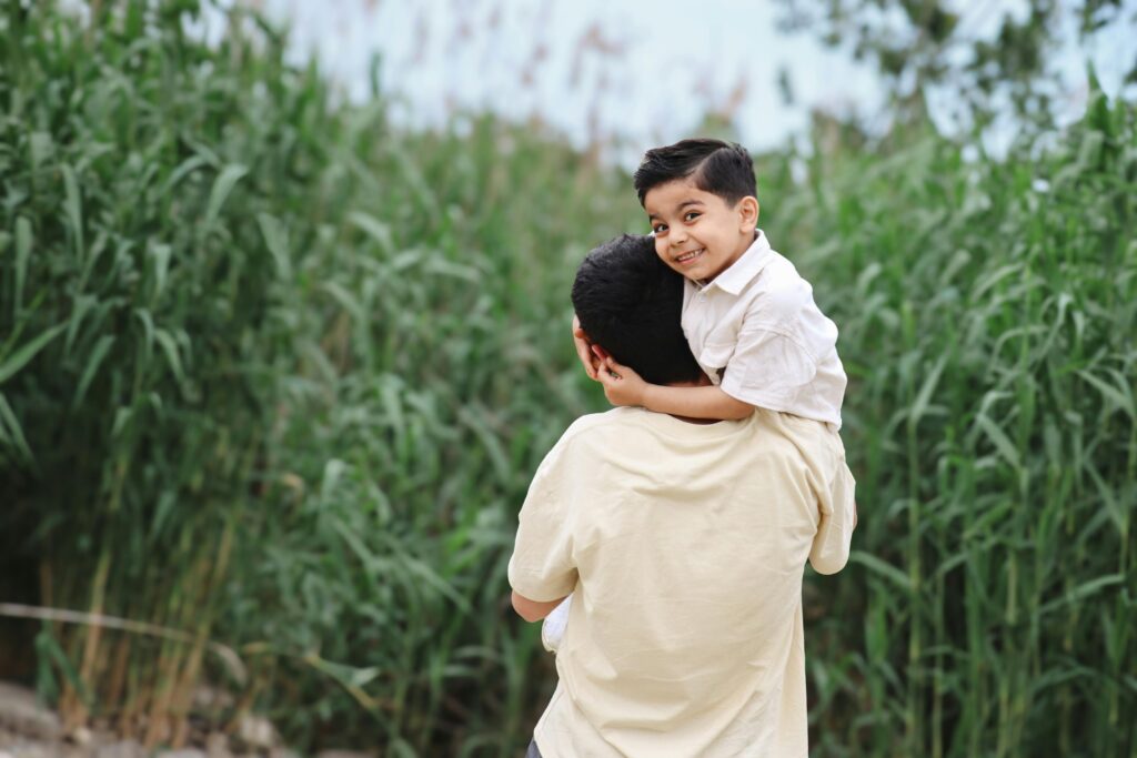 A joyful moment of a father carrying his smiling son on his shoulder in a green outdoor setting.